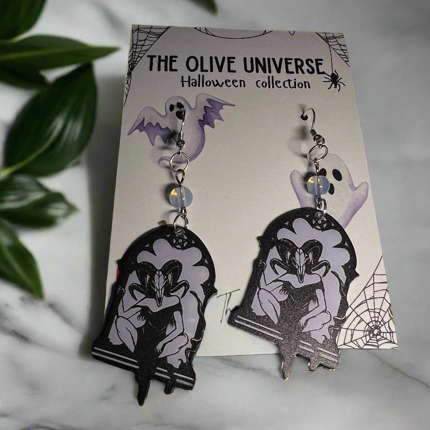 Halloween-themed earrings featuring occult style ram magic mask, occult symbolism, magic spell, women with ram mask on, opal beads on a branded card with spider webs, pumpkins, and ghosts.
