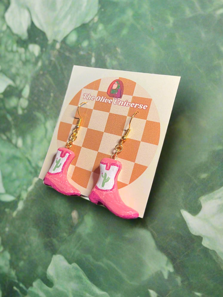 Pink cowboy boot earrings on a checkered card with 'The Olive Universe' branding, against a green leafy background.