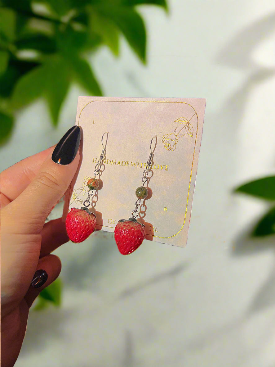 Strawberry-shaped earrings on a card held by a hand with dark nail polish, against a blurred background.