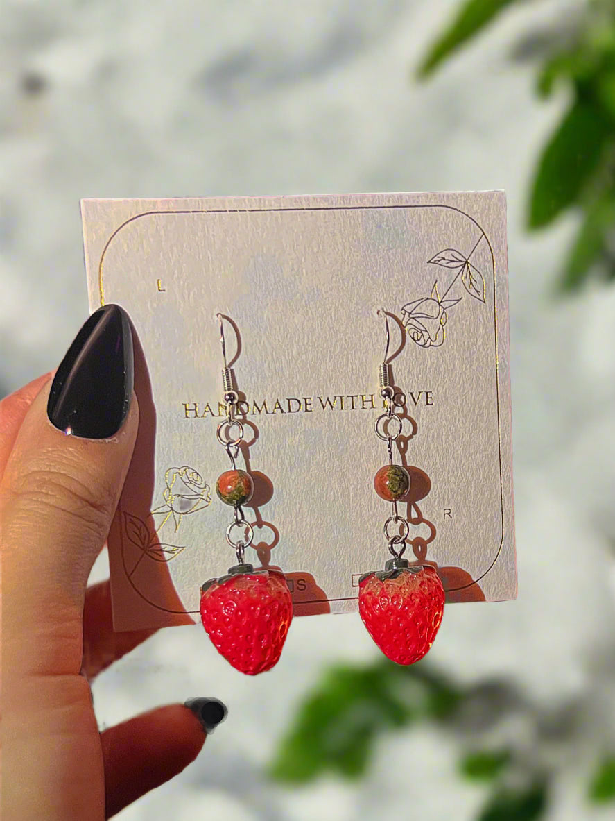 Strawberry-shaped earrings on a card held by a hand with black nail polish.