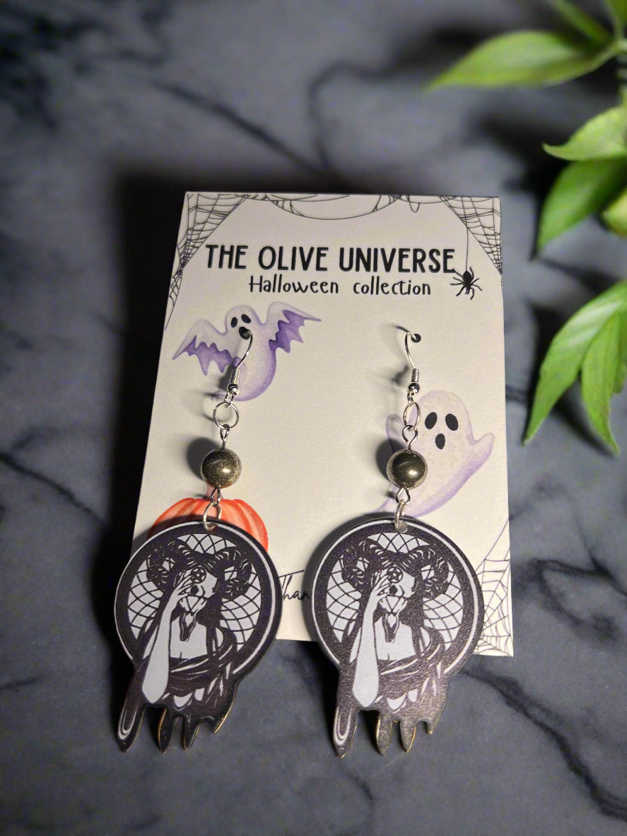 Halloween-themed earrings with dark witchy woman holding a mask. Features occult mask and symbolism. Spooky season earrings on a 'The Olive Universe' branded card with a ghost, pumpkin, and spiderweb background.