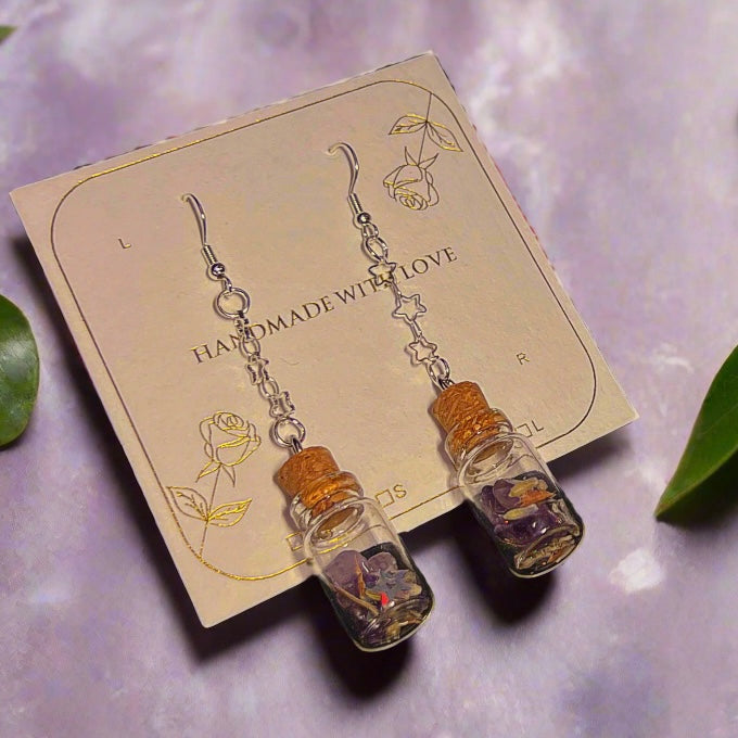 Pair of earrings glass bottles filled with amethyst, periwinkle, iridescent stars, and dangle from star-link chains. The witchy halloween spell jar earrings are on on a "handmade with love" card with floral designs