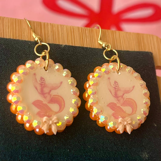 Handmade mermaid‑core earrings featuring a polymer‑clay merman portrait, 3D bow, orange pearls, rhinestones, and 18k gold‑plated hypoallergenic findings—whimsical, nostalgic, wearable art perfect for a statement look.