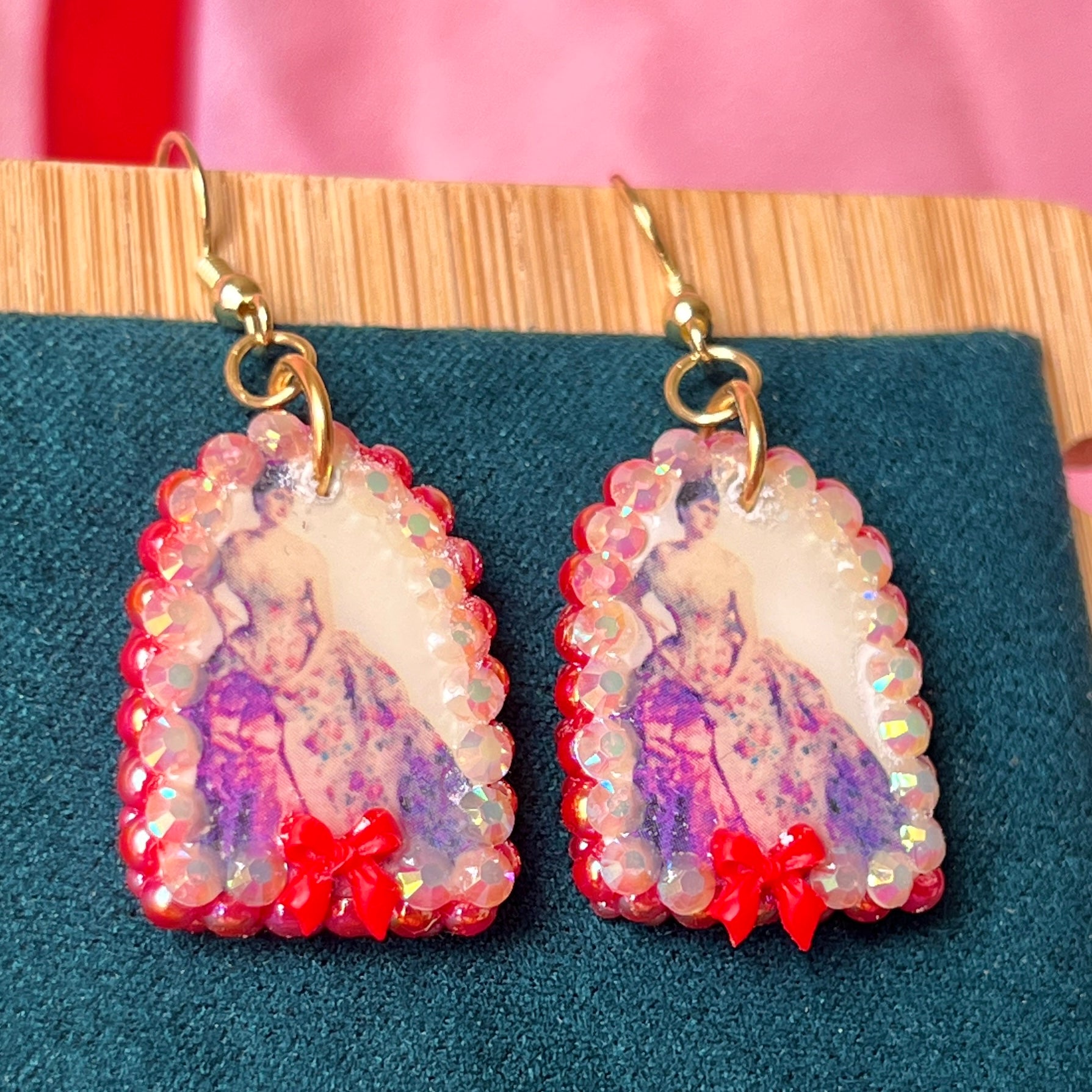 Alt text: A wearable art earring inspired by a Victorian romance portrait, featuring a polymer‑clay art print charm embellished with iridescent pearls, clear rhinestones, and a red coquette bow, hanging from gold‑plated hooks—perfect for museum‑core, renaissance‑core, and art‑lover styling.