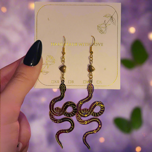 Snake-shaped oil slick iridescent earrings with gold hematite beads, gold accents, gold details, and hypoallergenic hooks on a card held by a hand against a purple blurred background
