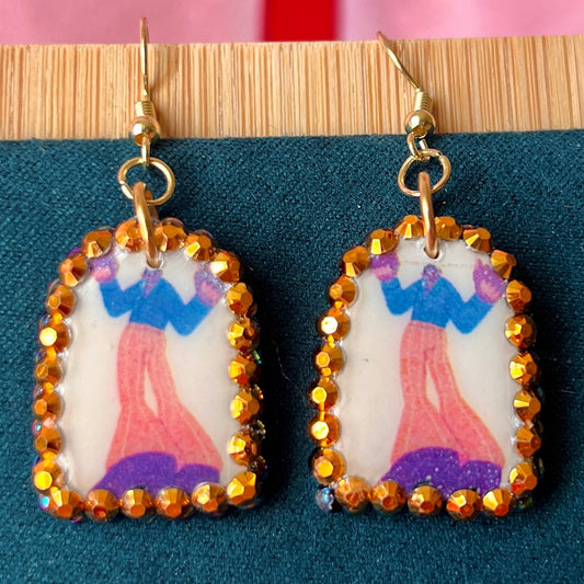 Handmade polymer‑clay arch earrings featuring a tall jazzy guy in orange pants and black boots, bedazzled with black, iridescent pearl and bright orange rhinestones, offering bold queer‑alt, punk‑glam, art‑kid style on 18k gold‑plated hooks.