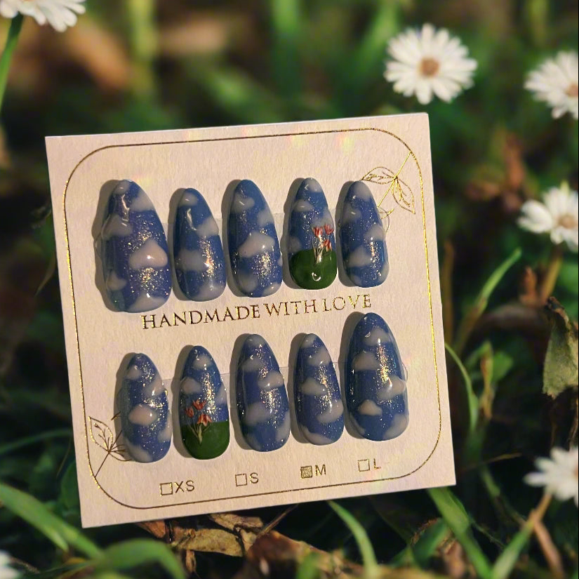 Set of blue and green nail art designs on a cardboard backing with 'Handmade with Love' text, placed on grass with flowers.