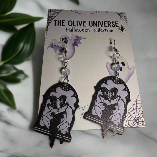 Halloween-themed earrings featuring occult style ram magic mask, occult symbolism, magic spell, women with ram mask on, opal beads on a branded card with spider webs, pumpkins, and ghosts.