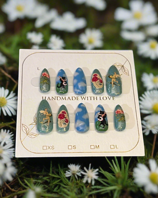Set of nail art designs on a cardboard backing with 'Handmade with Love' text, surrounded by white flowers. The nails have blue skys with 3D clouds, a field with a cat playing a stringed instrument, fairies, roses, and glitter.