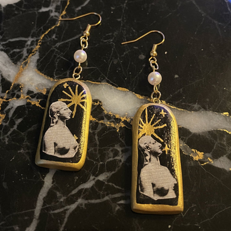 Pair of earrings with a design of a woman and sun, on a dark background.