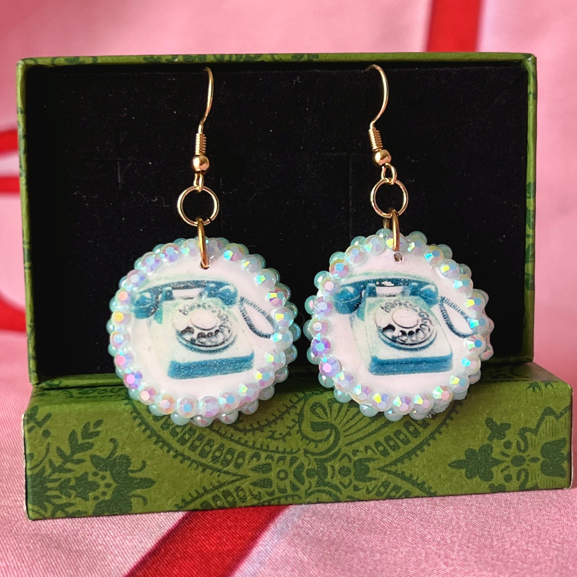 A handcrafted pair of iridescent teal dial‑up phone earrings made from polymer clay, featuring luminous pearl detailing, 18k gold‑plated hypoallergenic hooks, and a nostalgic Y2K, maximalist, statement‑jewelry vibe.