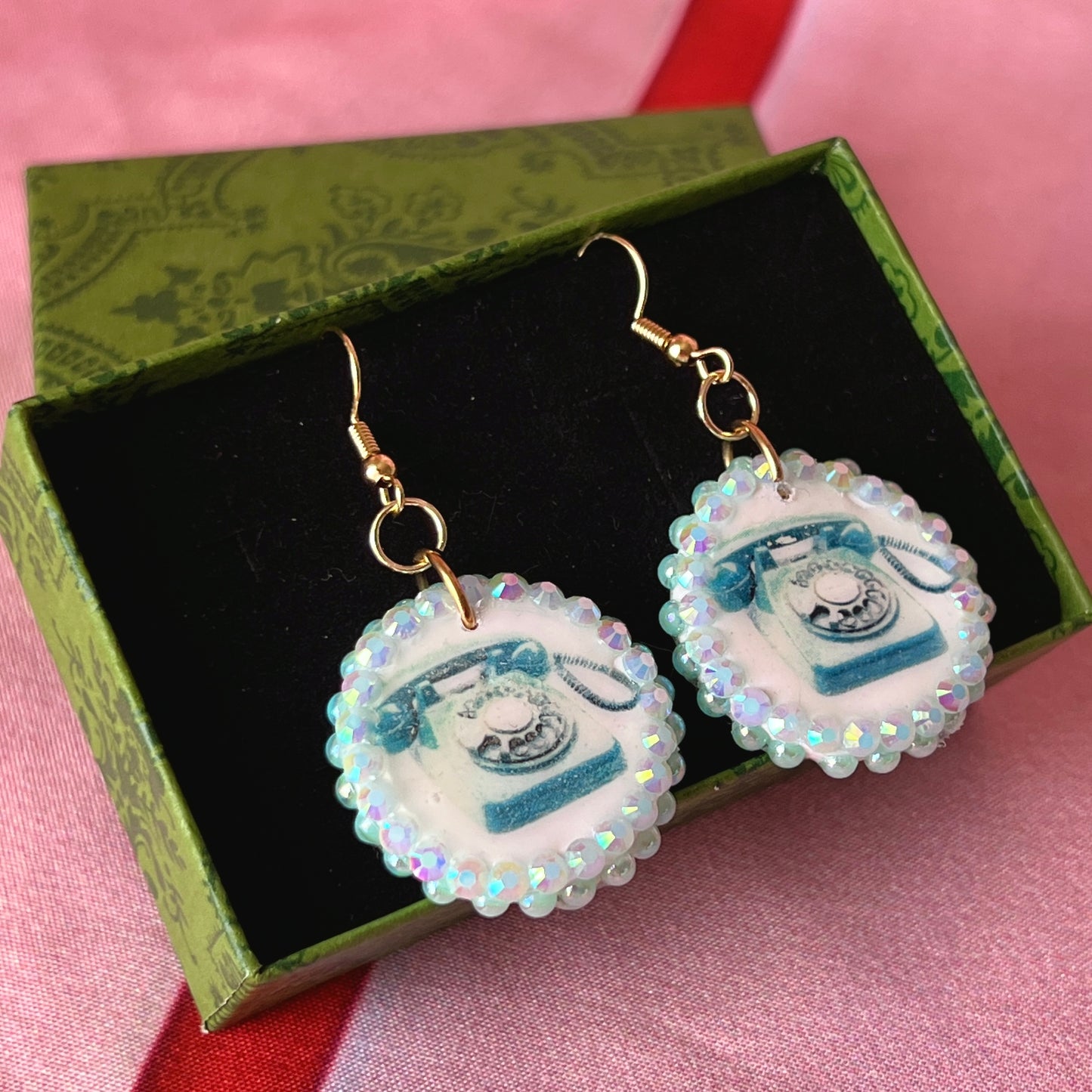 A handcrafted pair of iridescent teal dial‑up phone earrings made from polymer clay, featuring luminous pearl detailing, 18k gold‑plated hypoallergenic hooks, and a nostalgic Y2K, maximalist, statement‑jewelry vibe.