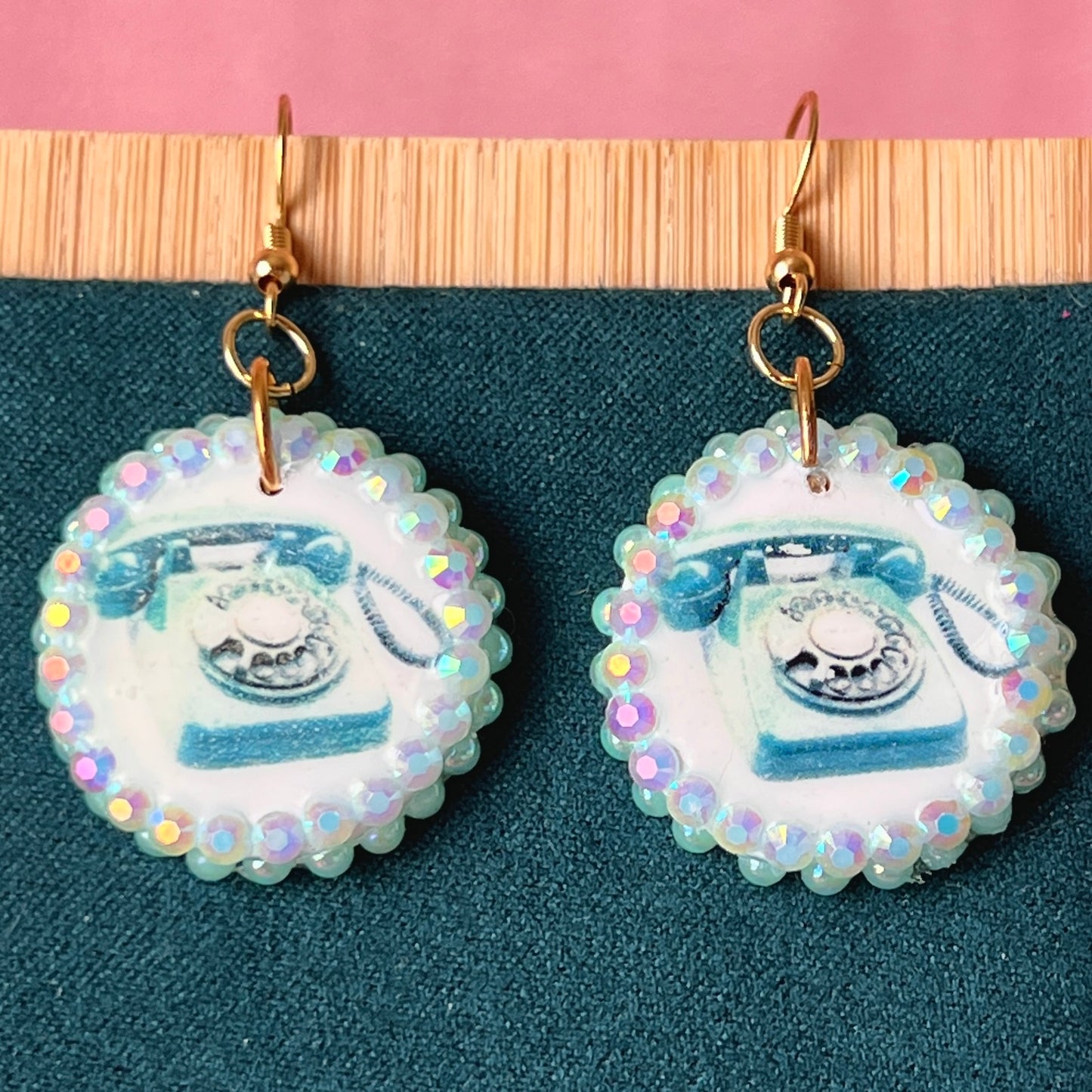 A handcrafted pair of iridescent teal dial‑up phone earrings made from polymer clay, featuring luminous pearl detailing, 18k gold‑plated hypoallergenic hooks, and a nostalgic Y2K, maximalist, statement‑jewelry vibe.