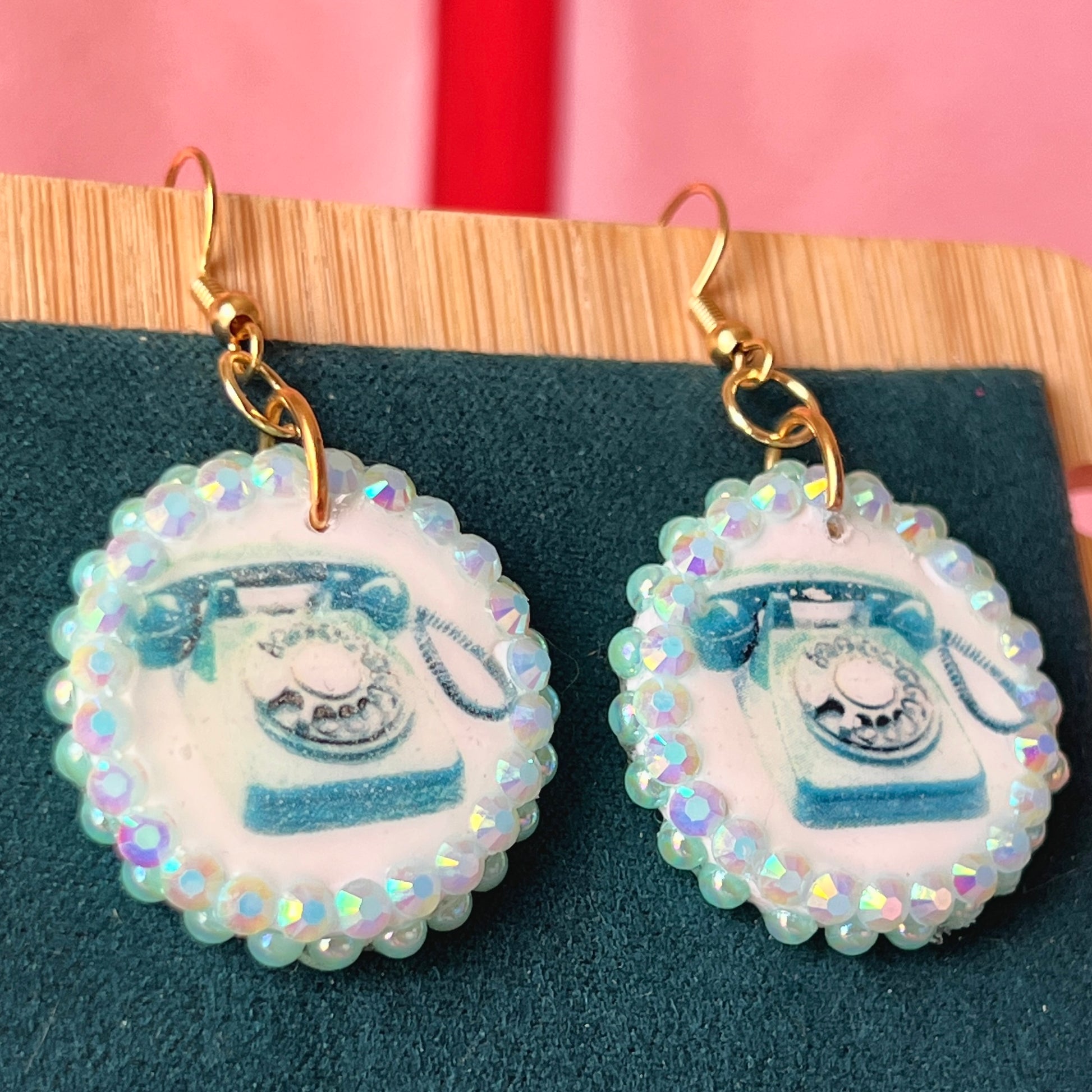 A handcrafted pair of iridescent teal dial‑up phone earrings made from polymer clay, featuring luminous pearl detailing, 18k gold‑plated hypoallergenic hooks, and a nostalgic Y2K, maximalist, statement‑jewelry vibe.