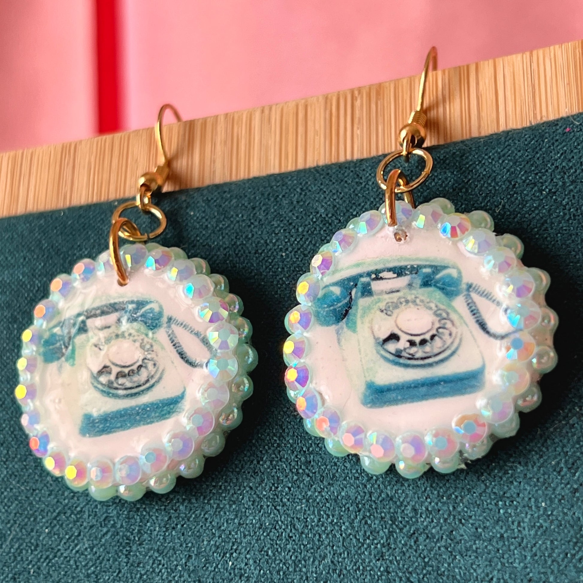 A handcrafted pair of iridescent teal dial‑up phone earrings made from polymer clay, featuring luminous pearl detailing, 18k gold‑plated hypoallergenic hooks, and a nostalgic Y2K, maximalist, statement‑jewelry vibe.
