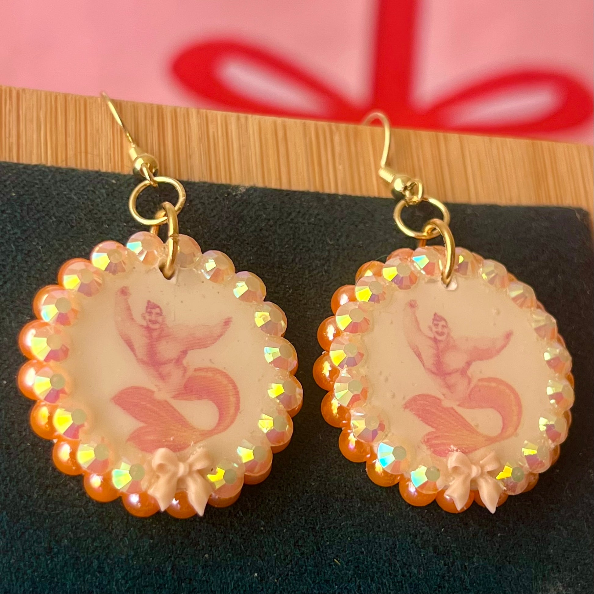 Handmade mermaid‑core earrings featuring a polymer‑clay merman portrait, 3D bow, orange pearls, rhinestones, and 18k gold‑plated hypoallergenic findings—whimsical, nostalgic, wearable art perfect for a statement look.