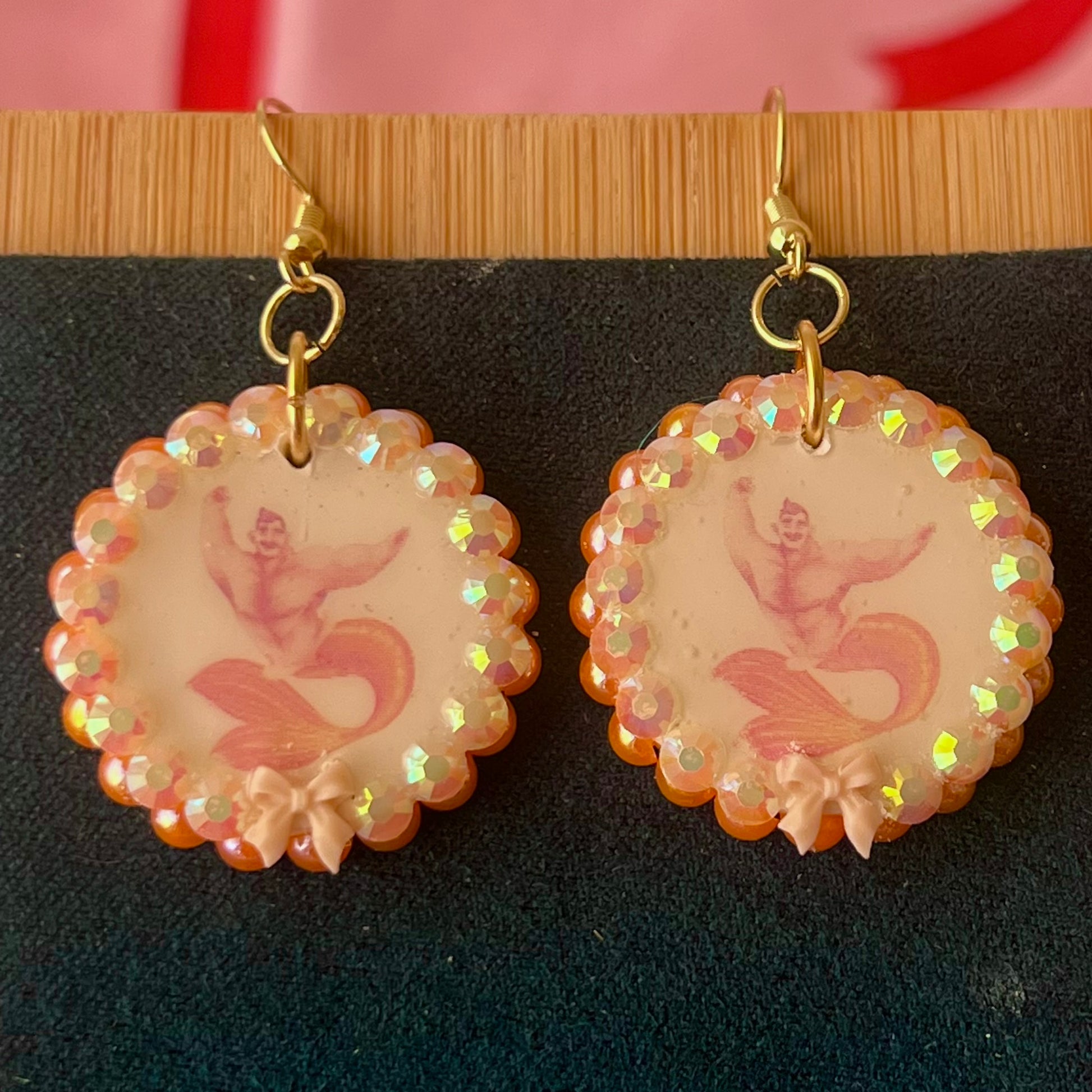 Handmade mermaid‑core earrings featuring a polymer‑clay merman portrait, 3D bow, orange pearls, rhinestones, and 18k gold‑plated hypoallergenic findings—whimsical, nostalgic, wearable art perfect for a statement look.