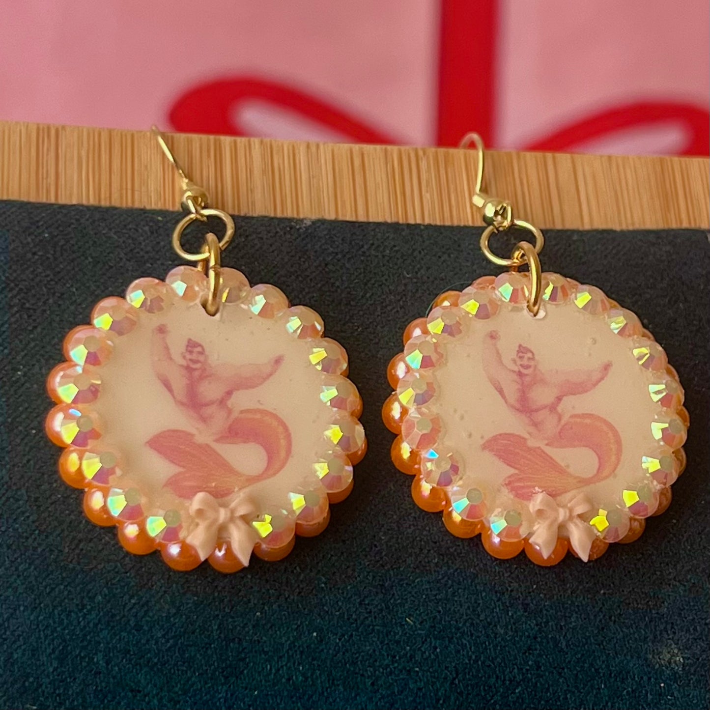 Handmade mermaid‑core earrings featuring a polymer‑clay merman portrait, 3D bow, orange pearls, rhinestones, and 18k gold‑plated hypoallergenic findings—whimsical, nostalgic, wearable art perfect for a statement look.