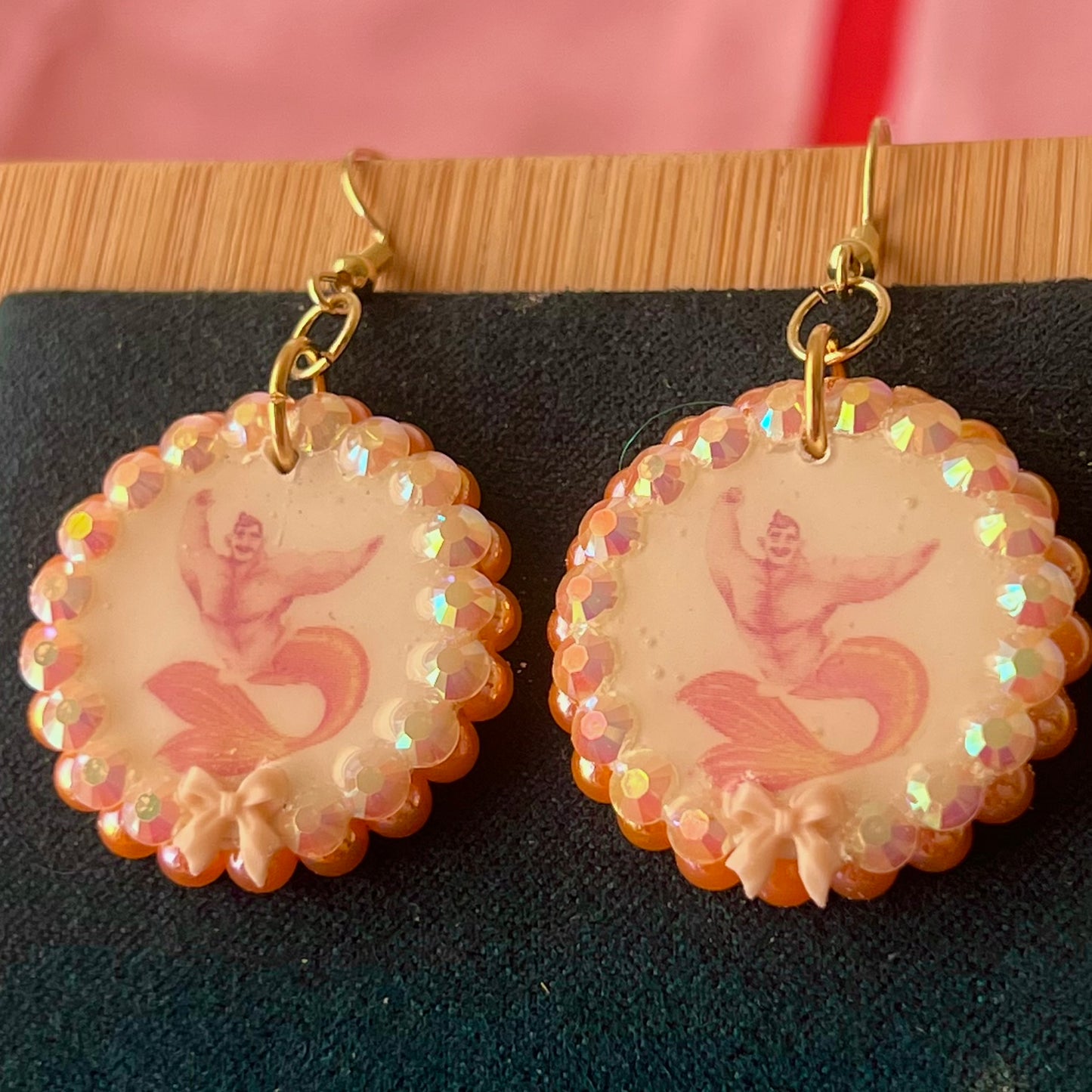 Handmade mermaid‑core earrings featuring a polymer‑clay merman portrait, 3D bow, orange pearls, rhinestones, and 18k gold‑plated hypoallergenic findings—whimsical, nostalgic, wearable art perfect for a statement look.