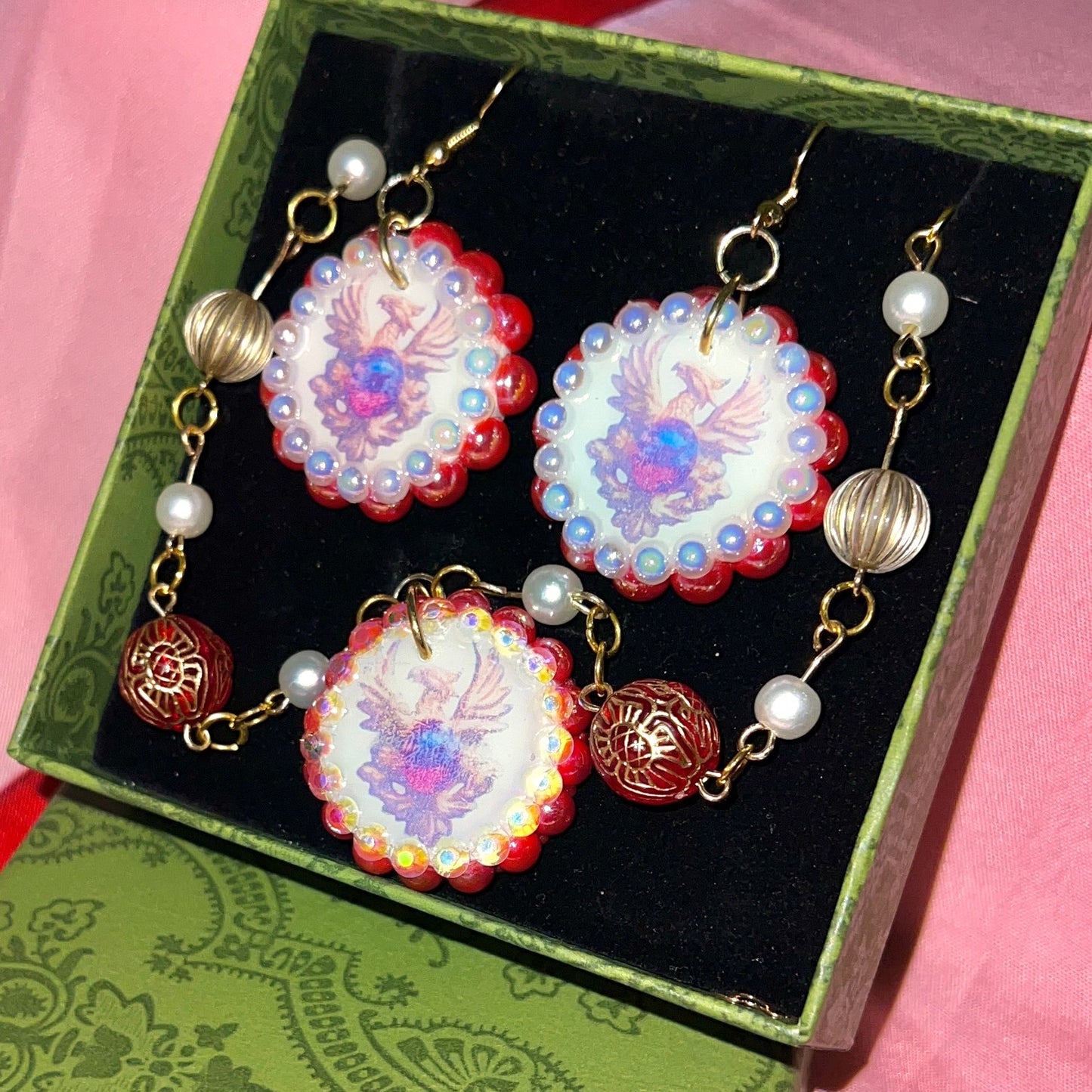 Alt text: A regal, medieval‑inspired pair of earrings featuring a golden phoenix sigil cradling a crimson gem, bedazzled with deep red and iridescent pearl‑like beads, perfect for fantasy, renaissance fair, and royal‑style looks.