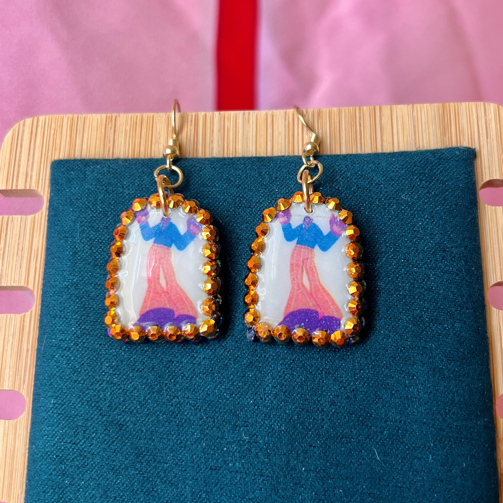 Handmade polymer‑clay arch earrings featuring a tall jazzy guy in orange pants and black boots, bedazzled with black, iridescent pearl and bright orange rhinestones, offering bold queer‑alt, punk‑glam, art‑kid style on 18k gold‑plated hooks.