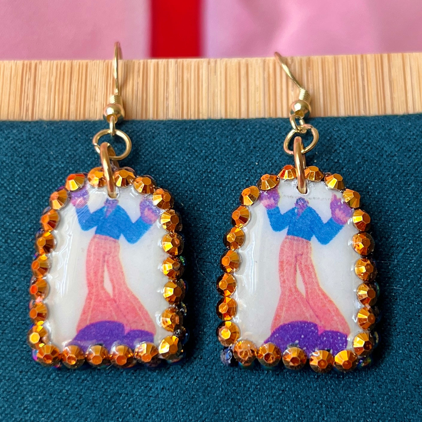 Handmade polymer‑clay arch earrings featuring a tall jazzy guy in orange pants and black boots, bedazzled with black, iridescent pearl and bright orange rhinestones, offering bold queer‑alt, punk‑glam, art‑kid style on 18k gold‑plated hooks.