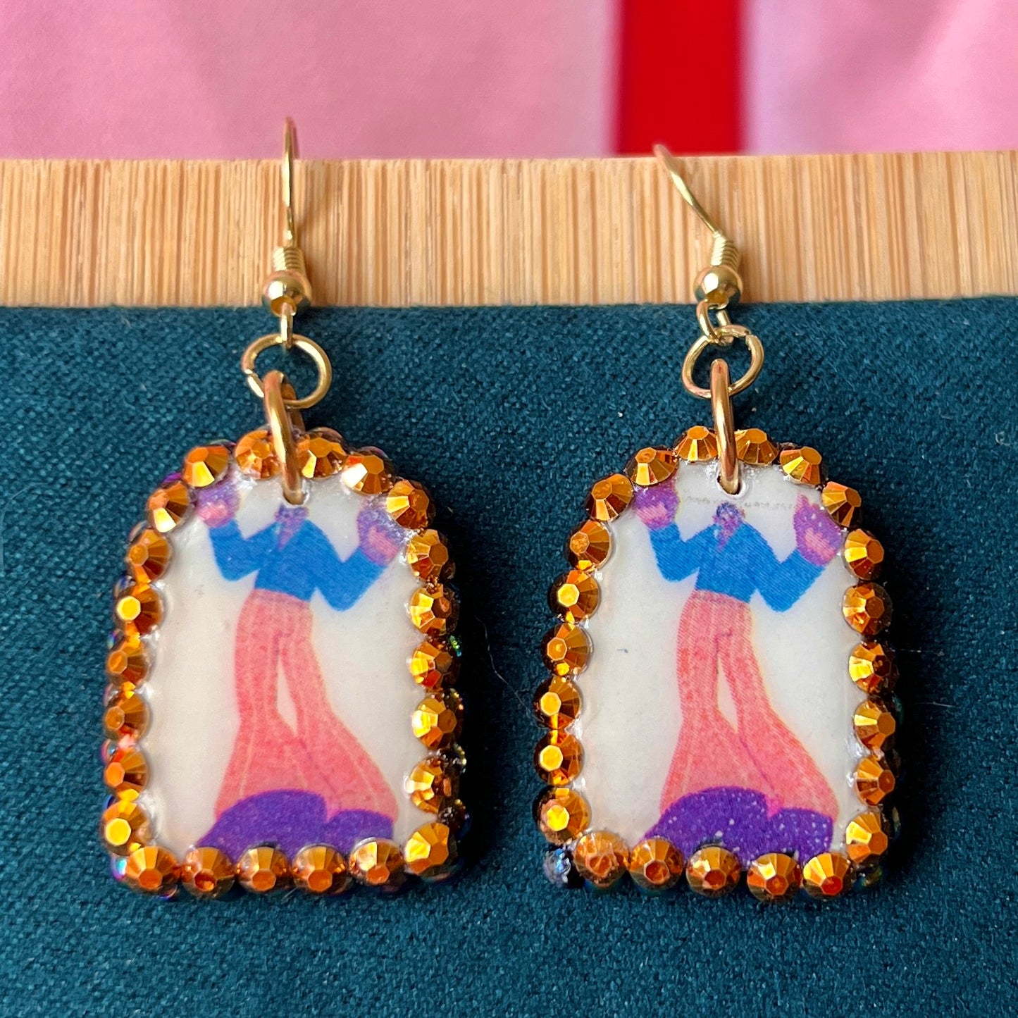Handmade polymer‑clay arch earrings featuring a tall jazzy guy in orange pants and black boots, bedazzled with black, iridescent pearl and bright orange rhinestones, offering bold queer‑alt, punk‑glam, art‑kid style on 18k gold‑plated hooks.