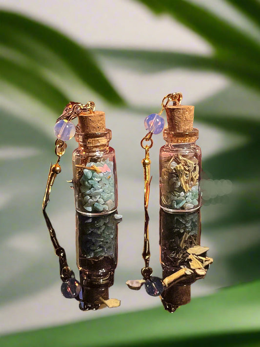 Pair of earrings resembling small glass bottles with cork lids, filled with green beads and decorative elements, on a blurred green leaf background.