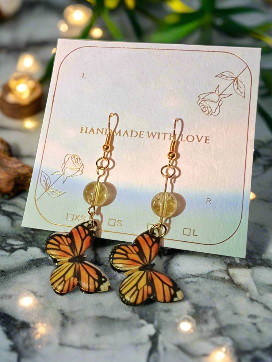 Butterfly earrings on a card with 'Handmade with Love' text, placed on a marble surface with decorative lights.