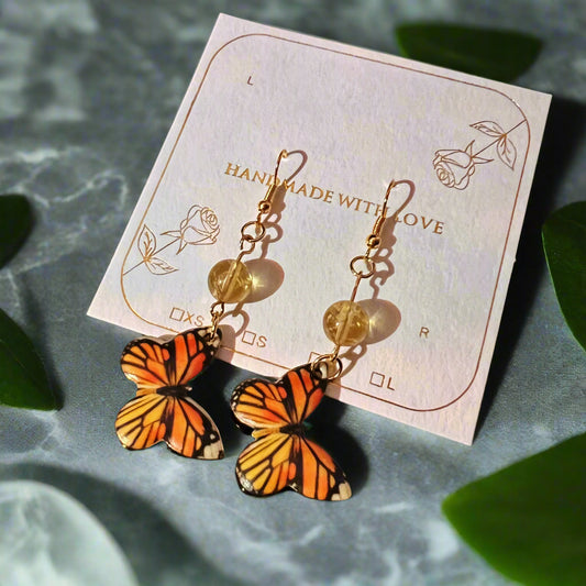 Butterfly-shaped earrings on a card with 'Handmade with Love' text, placed on a textured surface with green leaves.