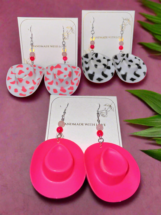 Pink hat-shaped earrings on a dark surface with other earrings and packaging in the background.