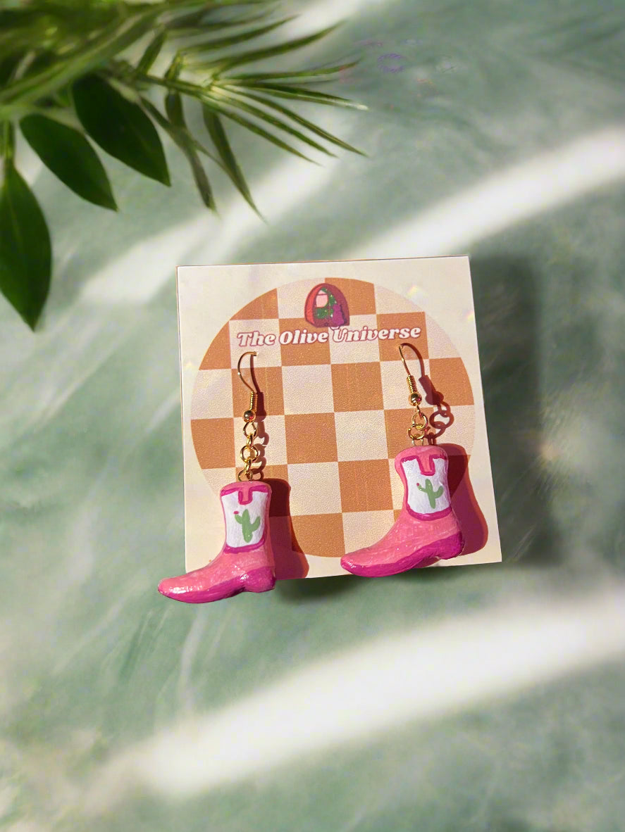 Pink cowboy boot earrings on a branded card with green leaves in the background