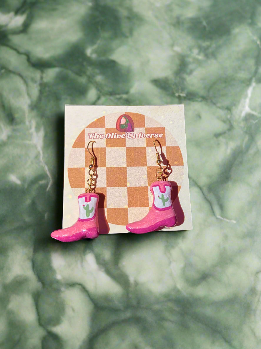 Pink boot-shaped earrings on a checkered card with 'The Olive Universe' branding against a green marble background.