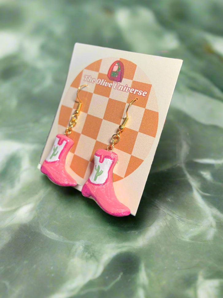 Pink cowboy boot earrings on a checkered card with 'The Olive Universe' branding, against a green water background.