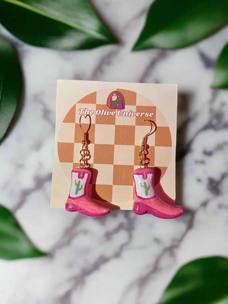 Pink cowboy boot earrings on a 'The Olive Universe' branded card with green leaves in the background