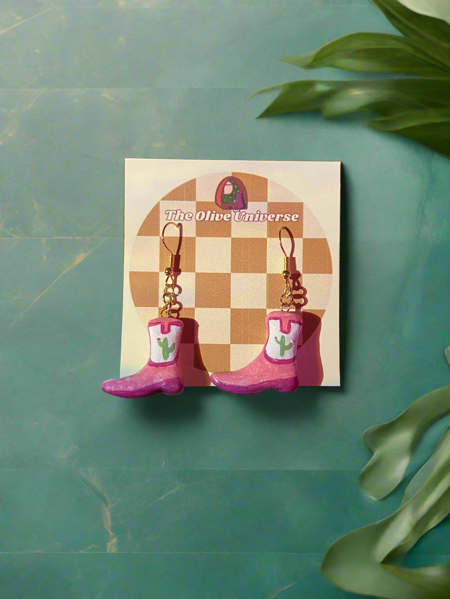 Pink cowboy boot earrings on a checkered card with 'The Olive Universe' branding against a green background.