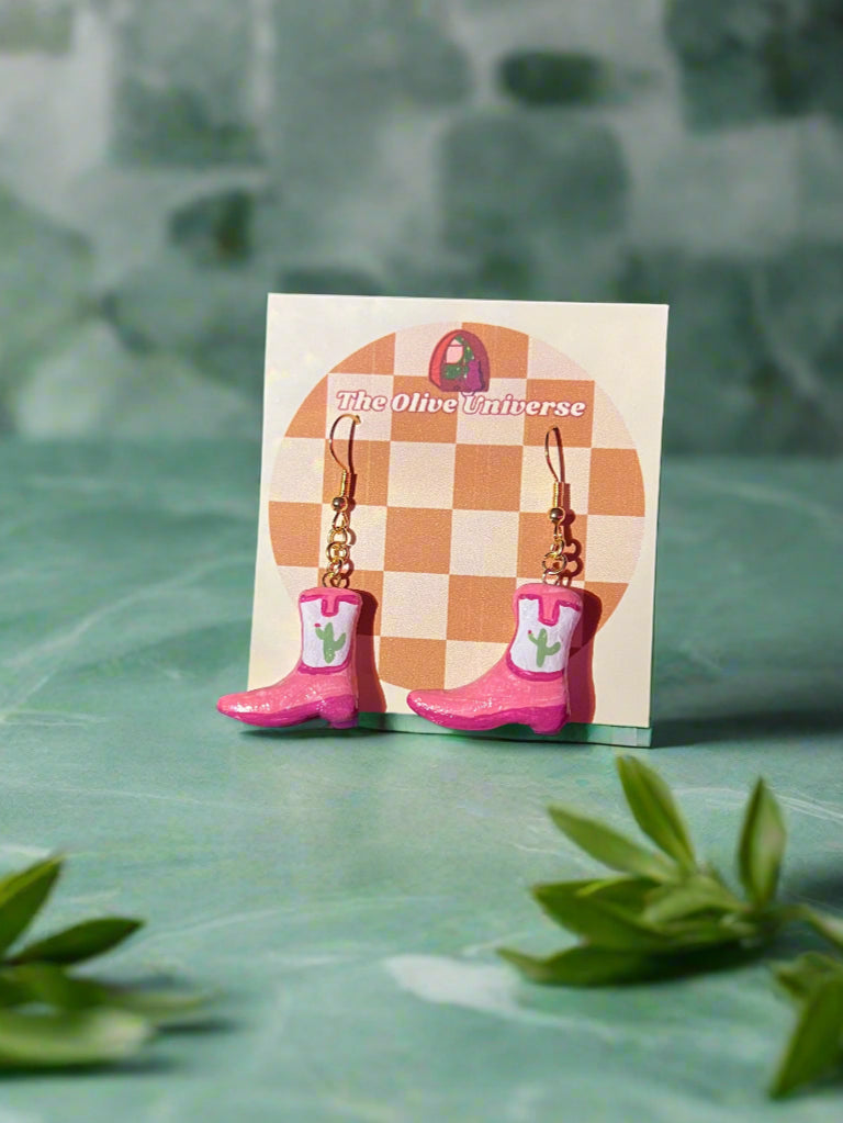 Pair of pink boot-shaped earrings on a checkered card with 'The Olive Universe' branding, against a dark background.