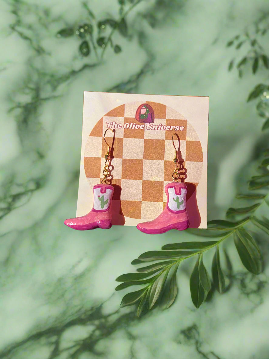 Pink boot-shaped earrings on a checkered card with 'The Olive Universe' branding, against a green leafy background.
