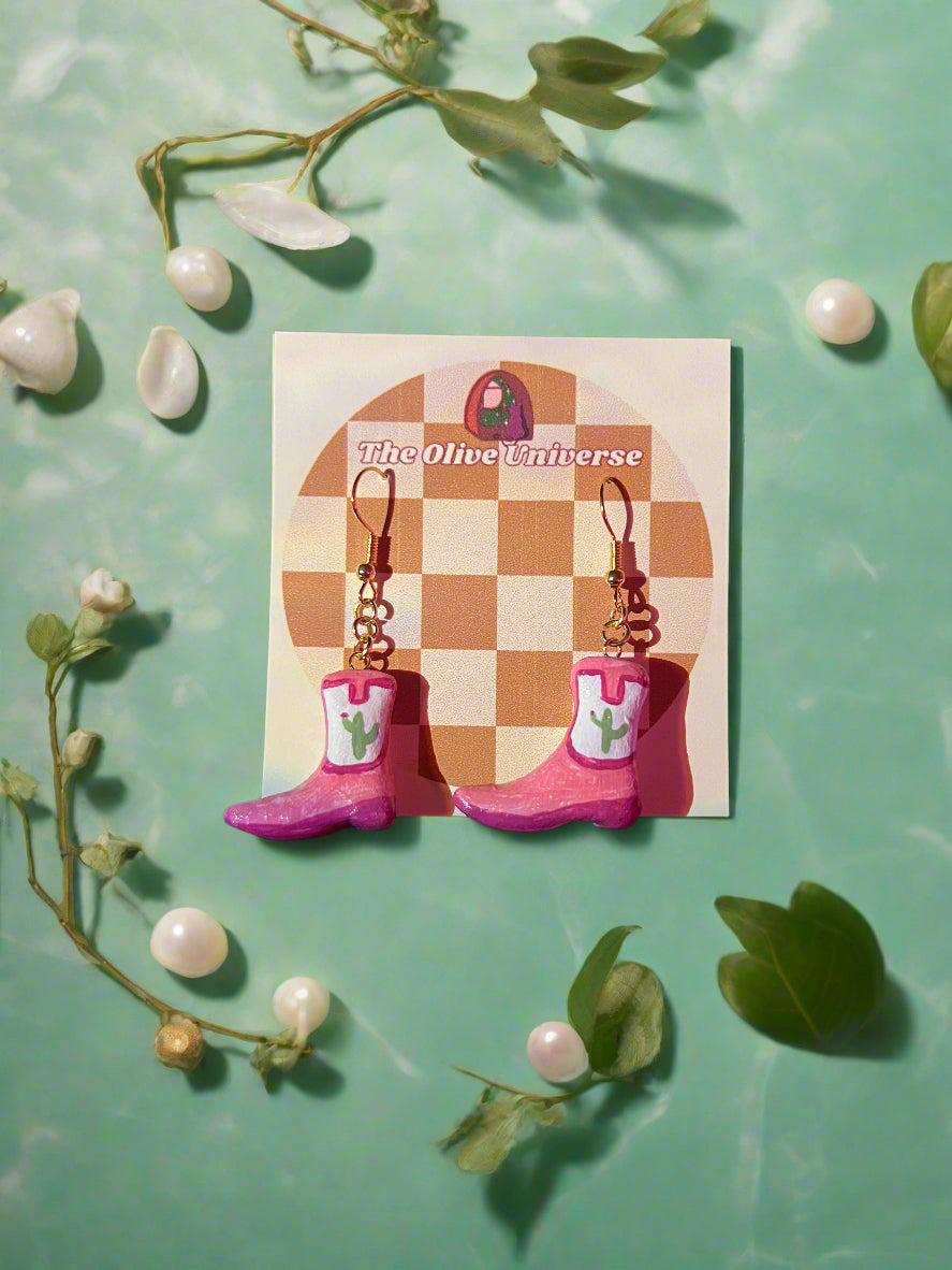 Pink cowboy boot earrings on a card with 'The Oliver Universe' branding, surrounded by green leaves and white flowers on a light green background.