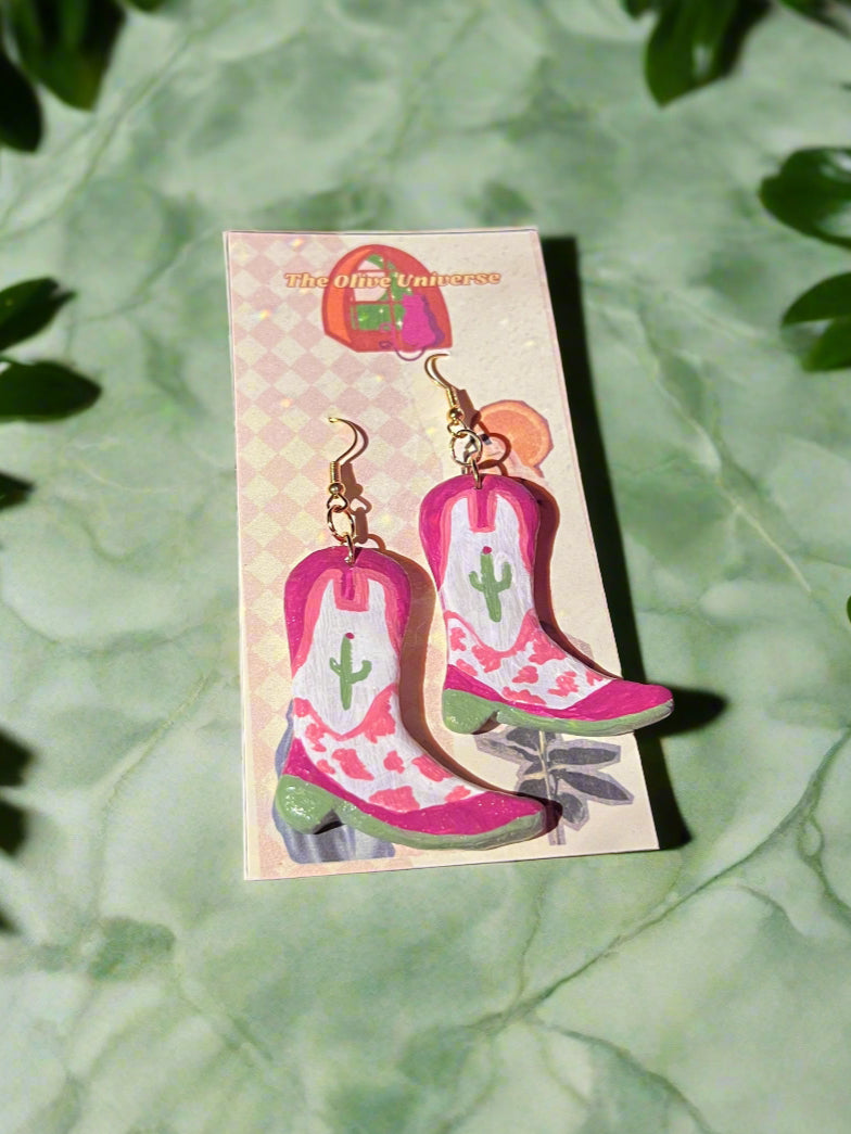 Pink cowboy boot earrings on a card with green leaves in the background