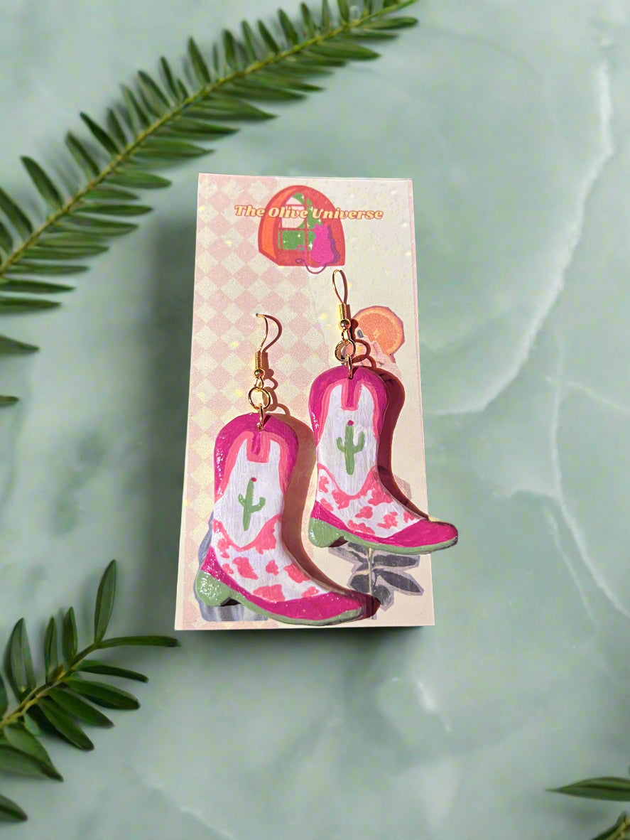 Pair of pink boot-shaped earrings on a card with a dark background