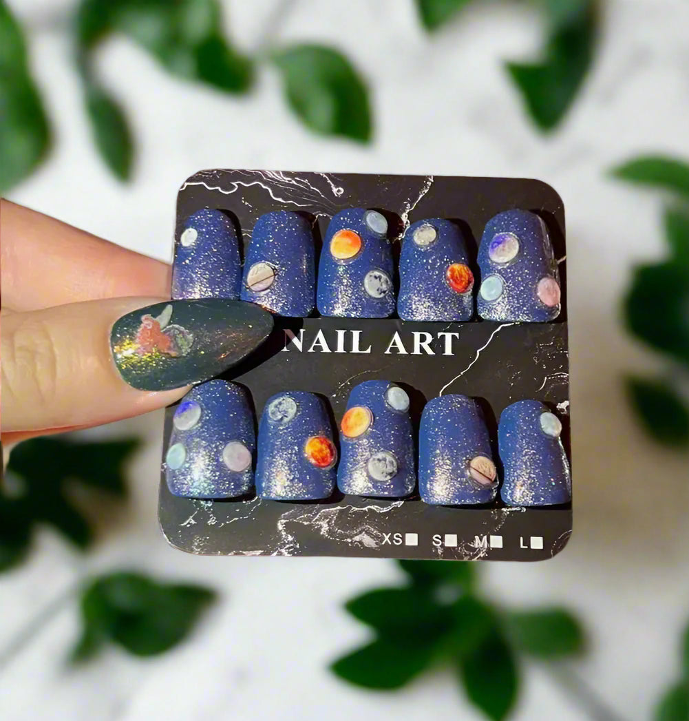 Nail art set with space-themed design held by a hand against a blurred green leaf background