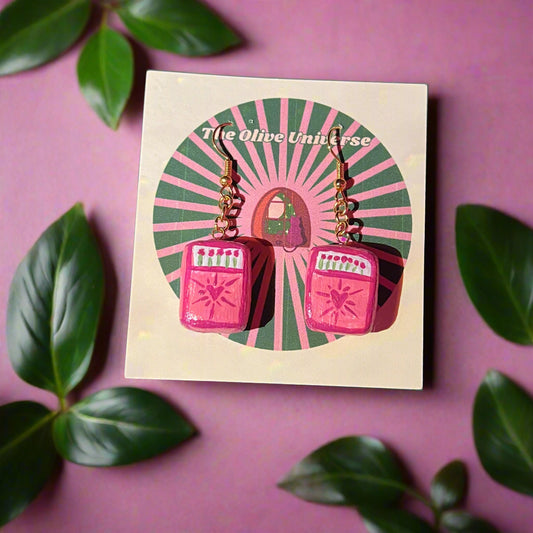 Pair of pink and gold earrings on a card with 'The Olive Universe' branding, surrounded by green leaves on a pink background.