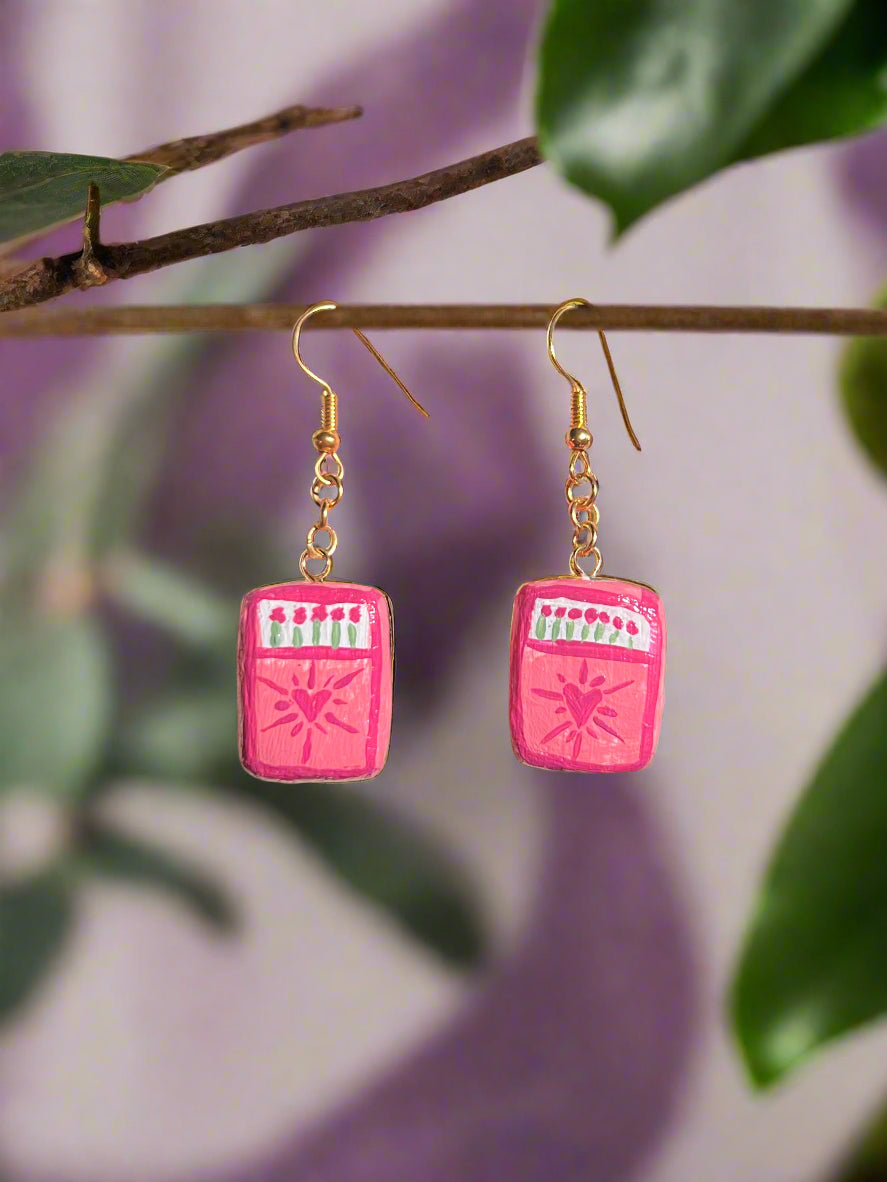 Pink earrings with heart designs hanging on a branch with leaves.