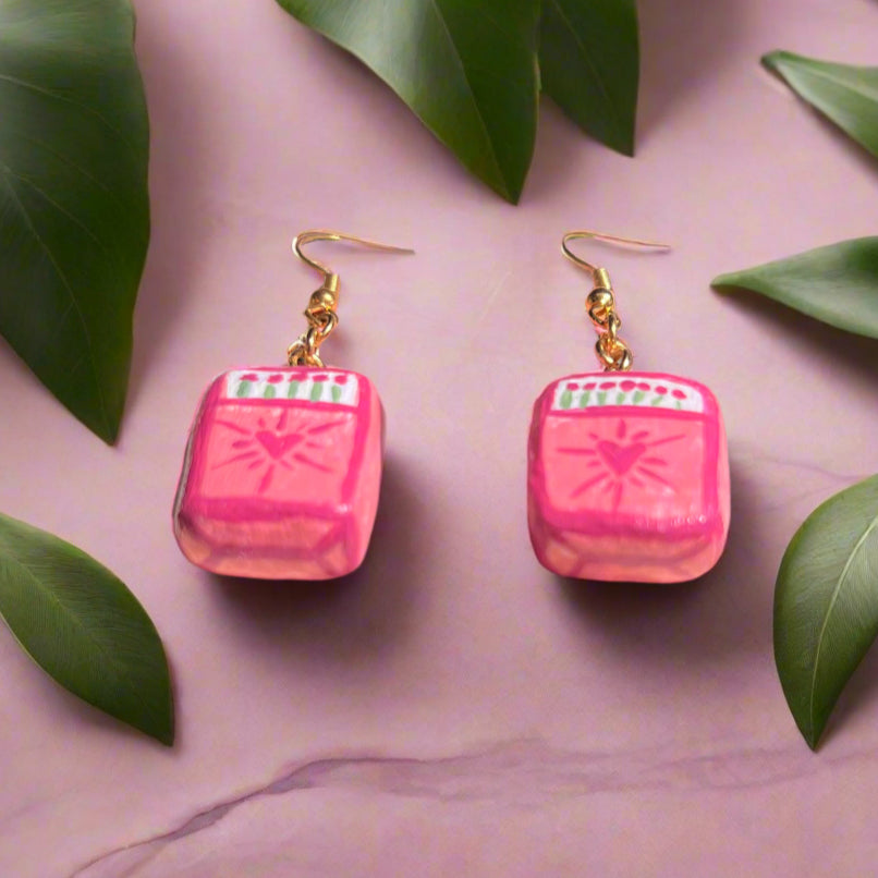 Pink square earrings with gold hooks on a pink background with green leaves.