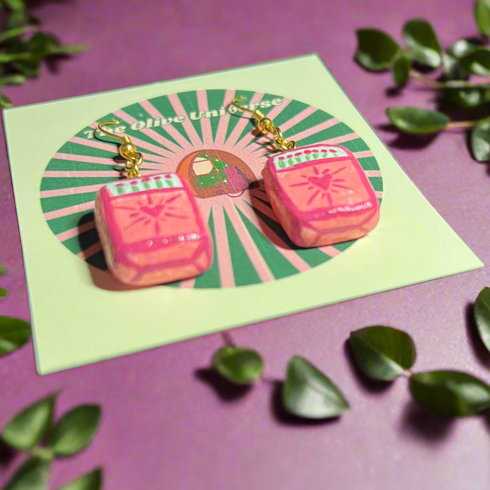 Pink earrings on a green card with a purple background and green leaves.