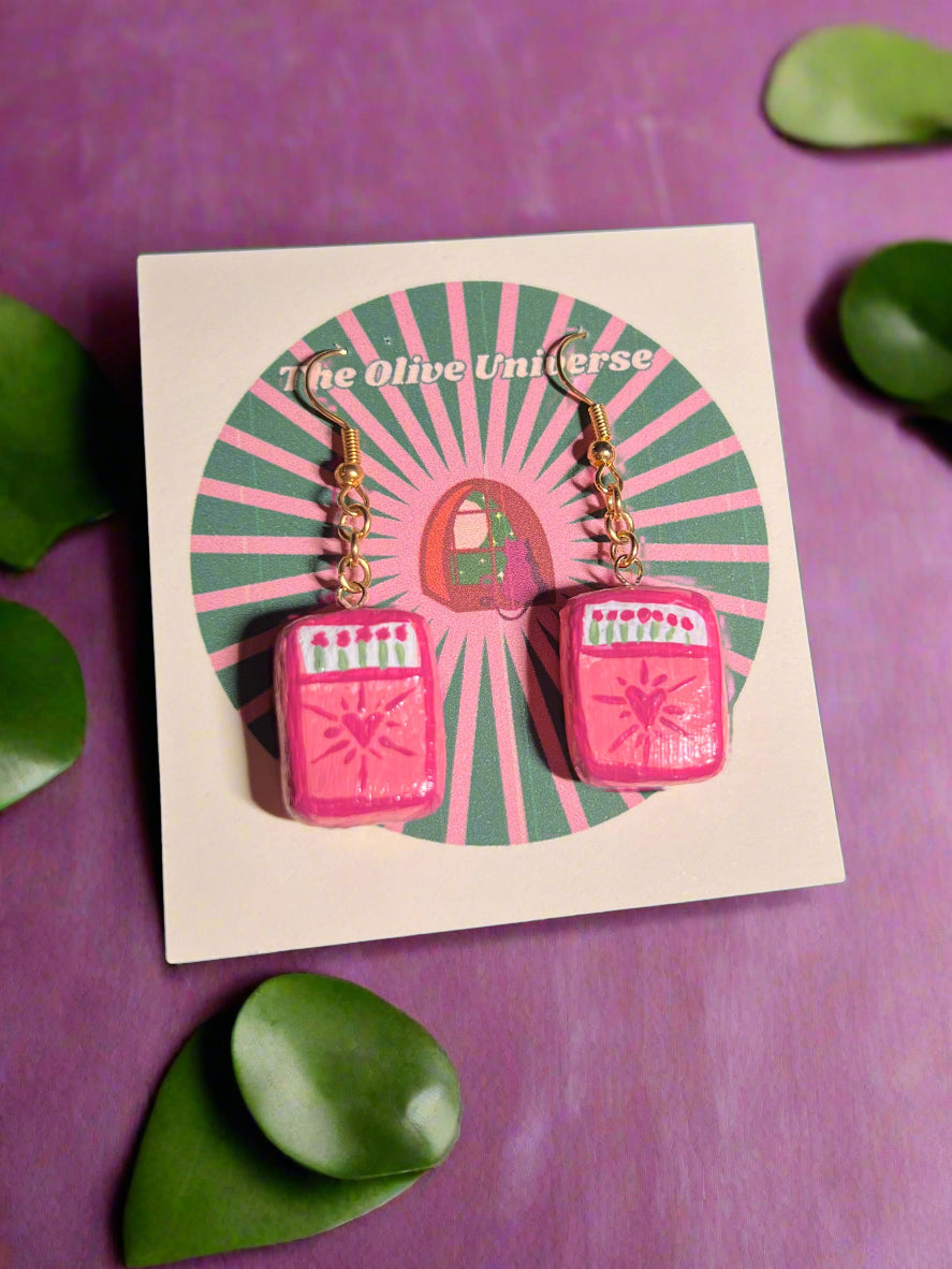 Pink earrings on a card with 'The Olive Universe' branding, surrounded by green leaves on a purple background.