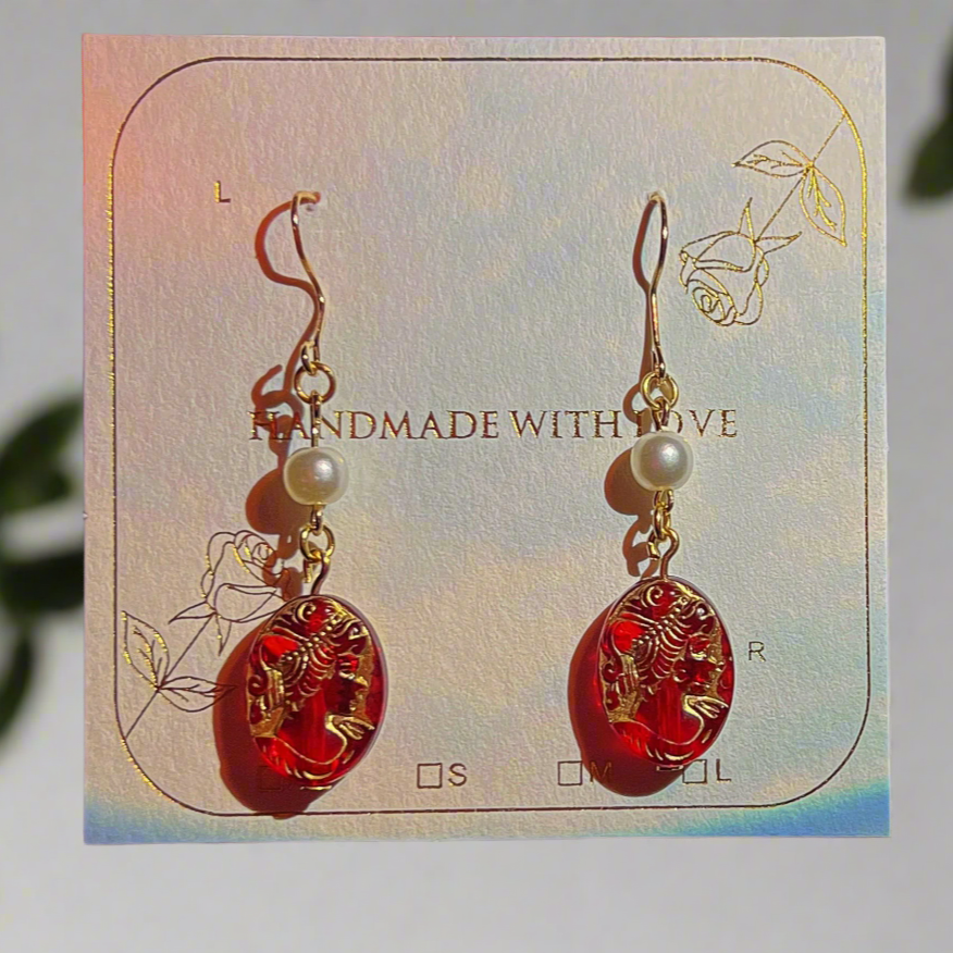 Red and gold earrings on a card with 'Handmade with Love' text, placed on a reflective surface.