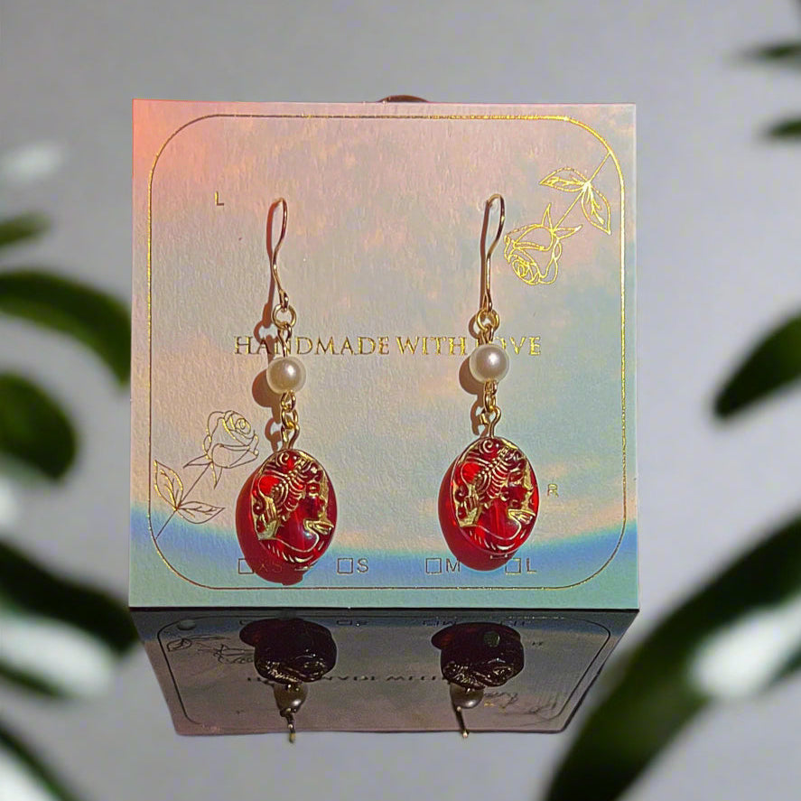 Red and gold earrings on a card with a candle and decorative items in the background