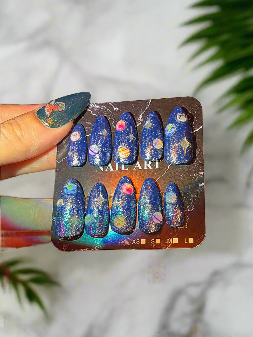 Set of nail art with space-themed design on a display card, held by a hand with blue nail polish.
